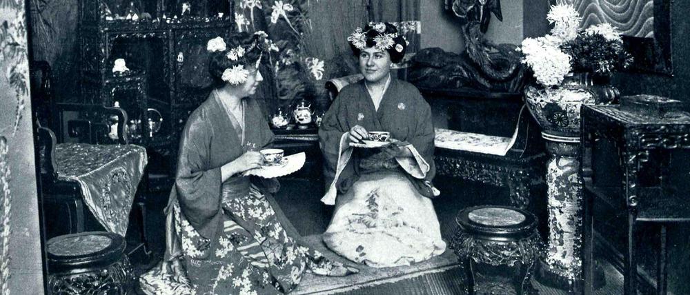 Zwei Damen sitzen bekleidet in Kimonos in einem japanisch eingerichteten Zimmer und halten Teetassen in der Hand.