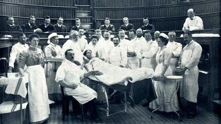 Der Chirurg Ernst von Bergmann mit grauem Bart steht im Operationssaal am Kopfende einer liegenden Patientin. Der Arzt ist umgeben von Assistenzärzten, Pflegern und Krankenschwestern.