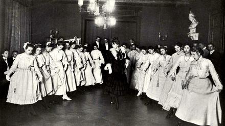 In zwei Reihen haben sich Mädchen in weißen Kleidern zum Tanzunterricht aufgestellt, in der Mitte steht die Tanzlehrerin. 