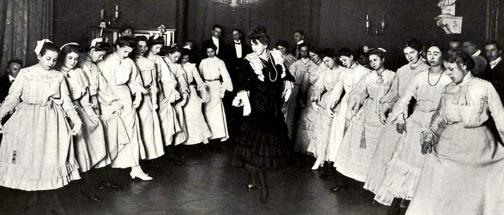 In zwei Reihen haben sich Mädchen in weißen Kleidern zum Tanzunterricht aufgestellt, in der Mitte steht die Tanzlehrerin. 