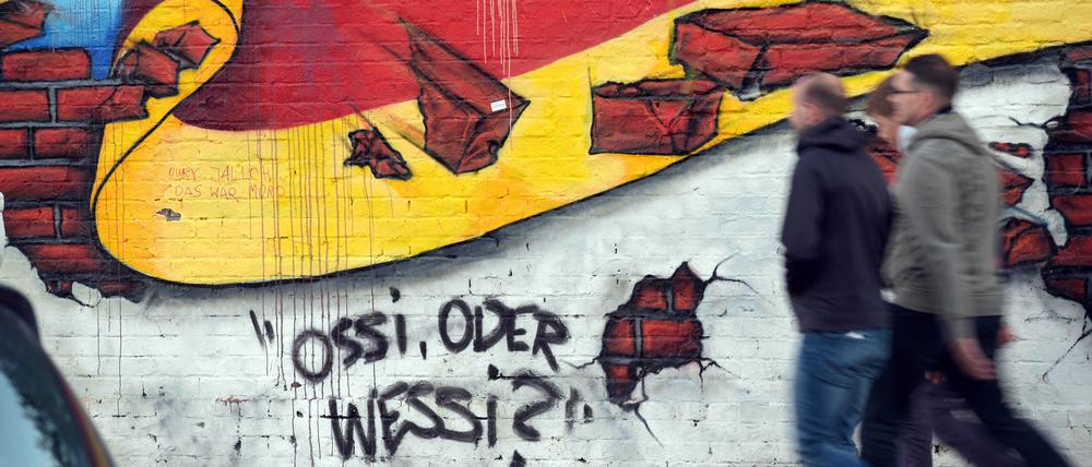 Passanten gehen an einem Wandbild von Caspar Kirchner mit der deutschen Nationalflagge und dem Schriftzug „Ossi oder Wessi?“ vorbei (Symbolbild).