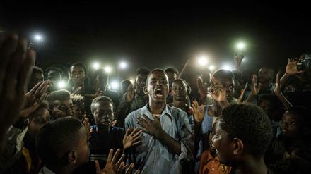 Das Bild zeigt, wie ein junger Sudaner umgeben von anderen Demonstranten im Dunklen inbrünstig ein Gedicht rezitiert - erhellt wird das Bild nur von Handylichtern der Umstehenden.