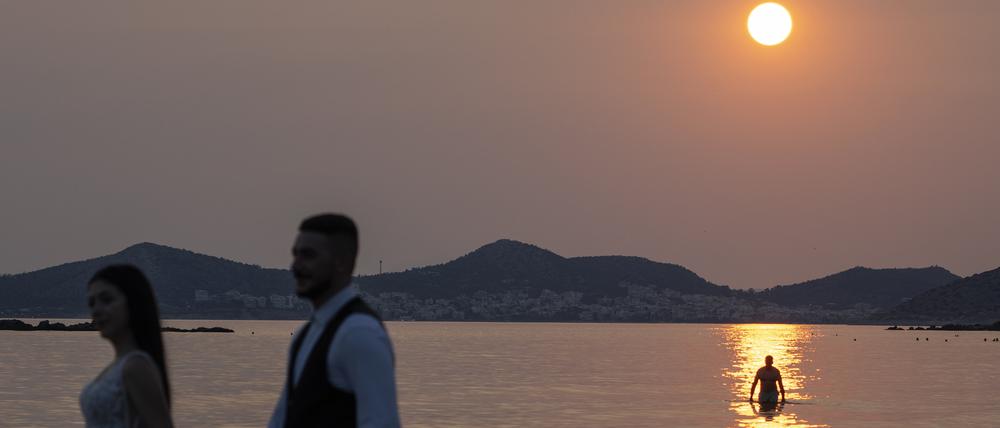 Ein frisch verheiratetes Paar posiert bei Sonnenuntergang für Fotos, während ein Mann ein Bad im Wasser in Lagonisi südöstlich von Athen nimmt.