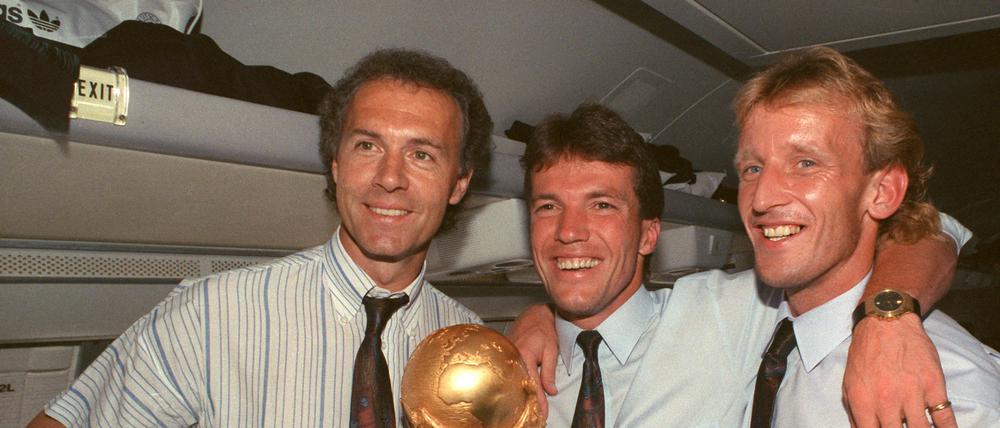 Im Flugzeug beim Rückflug von Rom nach Frankfurt präsentieren DFB-Teamchef Franz Beckenbauer (l), Kapitän und Mittelfeldspieler Lothar Matthäus (M) und Abwehrspieler Andreas Brehme strahlend den zwei Tage zuvor gewonnenen WM-Pokal. 
