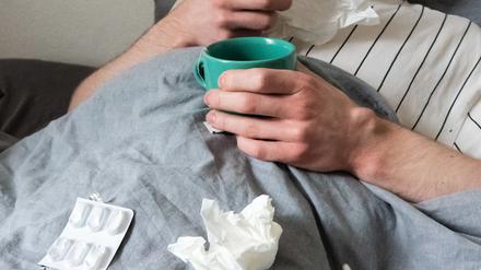 Ein Mann liegt mit Taschentüchern, Teetasse, Nasenspray und Tabletten im Bett (gestellte Szene). (Symbolbild)