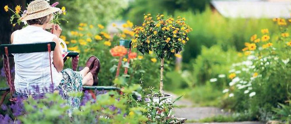 Eine Frau mit Sonnenhut sitzt kaffeetrinkend auf einem schlichten Holzstuhl in einem idyllischen Garten