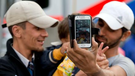 Zwei junge Männer mit einem kleinen Jungen auf dem Arm halten ein Smartphone in die Kamera.
