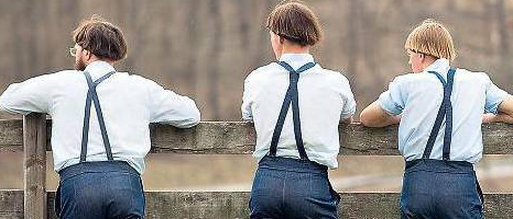 Drei erwachsene Amish und ein Kind stehen mit dem Rücken zum Betrachter an einem Zaun.