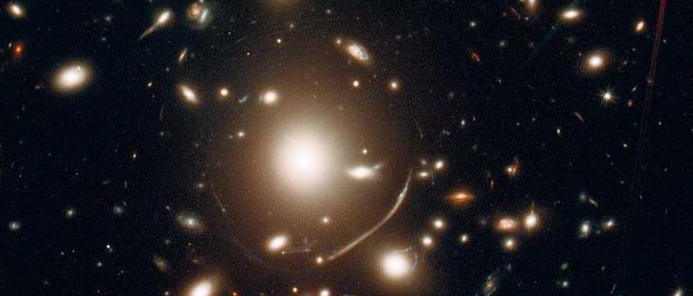 Kosmische Lichtspiele. Der Galaxienhaufen „Abell 383“ wirkt wie eine Linse, die das Licht weit entfernter Galaxien bündelt und verstärkt. Die Bilder sind jedoch verzerrt. Das zeigt sich besonders an den leuchtenden Bögen um die elliptische Galaxie (unten links). 