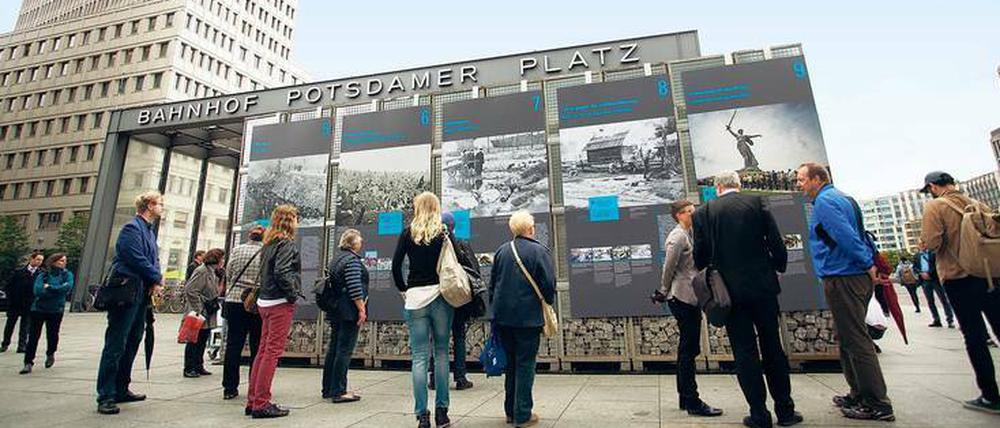 Auf einem von Hochhäusern gesäumten Platz und dem Eingang des Bahnhofs Potsdamer Platz steht eine Freiluft-Ausstellung, davor stehen Menschen, die die Tafeln der Ausstellung lesen.
