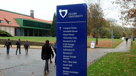 Studierende laufen bei herbstlichem Regenwetter über einen Campus, im Vordergrund ein Hinweisschild zu Unigebäuden.