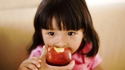 Gesunder Genuss. Ein kleines Mädchen beißt in einen Apfel, den ihr die Hand einer Erwachsenen hinhält.