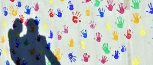 Auf einer Betonwand mit bunten Abdrücken von Kinderhänden ist ein Mann mit Kind auf dem Arm und an der Hand zu sehen.