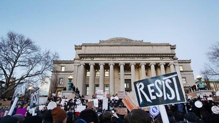 Vor einem historischen Universitätsgebäude stehen junge Menschen, die Plakate mit Aufschriften wie "Resist" hochhalten.