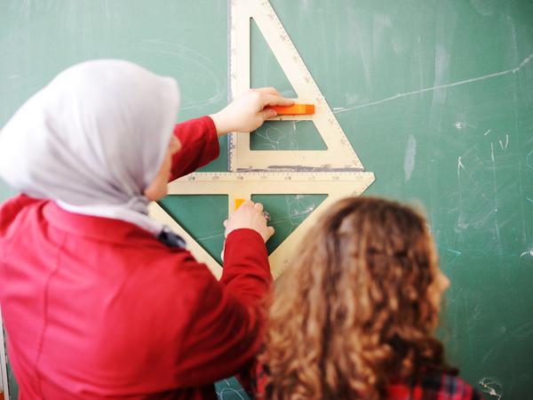 Eine Lehrerin mit Kopftuch steht mit einer Schülerin vor der Tafel. 
