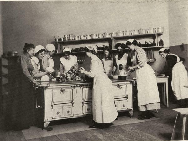 Das Bild stammt aus einem1912 erschienenen Band und zeigt Teilnehmerinnen eines dreimonatigen Koch-und Backkurses dieser 1866 gegründeten Hauswirtschaftsschule. Links am Herd steht die damalige Kochlehrerin und Autorin Elise Hannemann. 