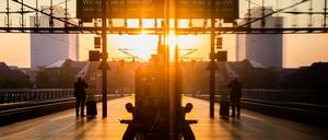 Fahrgäste sind am Hauptbahnhof bei Sonnenaufgang auf dem Bahnsteig zu sehen, wobei sich die Szenerie in einer Vitrine spiegelt.
