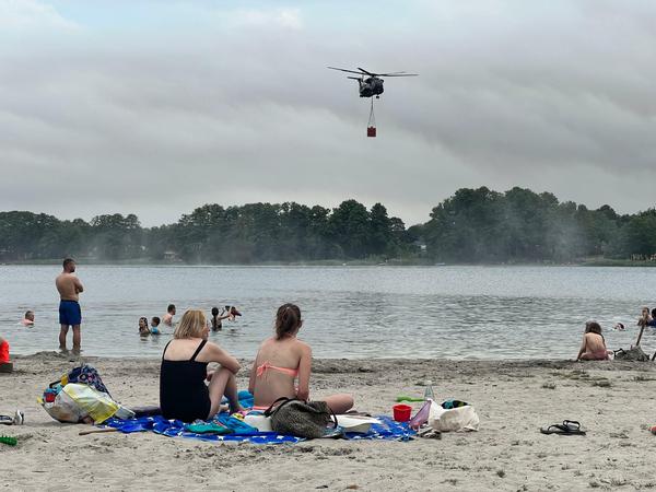 Bizzare Szenerie. Badegäste hocken am Strand des Seddiner Sees, während ein Löschhubschrauber der Bundeswehr Wasser schöpft.