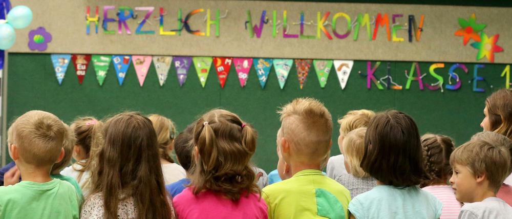 Erstklässler in einer Grundschule sitzen auf einer Bank mit dem Rücken zur Kamera. Da drüber ein "Herzlich Willkommen"-Transparent.