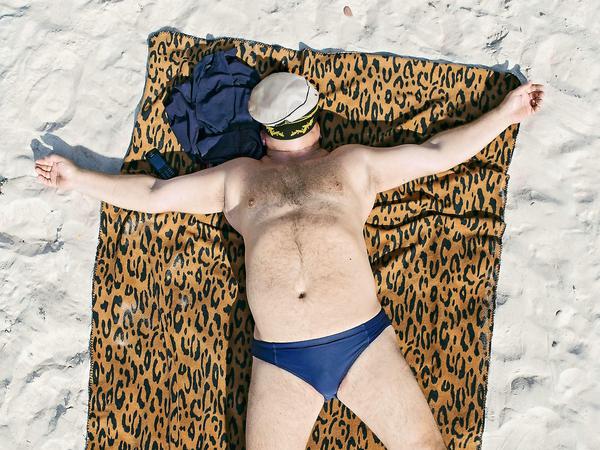 Ein Mann in knapper, blauer Badehose liegt ausgestreckt auf einem Strandtuch mit Leoparden-Muster. Eine Kapitänsmütze liegt auf seinem Gesicht.