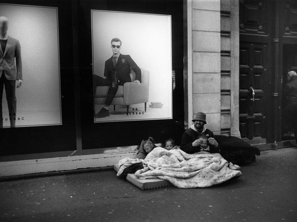 Paris 2014: Ein Obdachloser sitzt mit seinen drei Kindern auf einer Matratze, während im Hintergrund die Werbung eines Herrenausstatters leuchtet