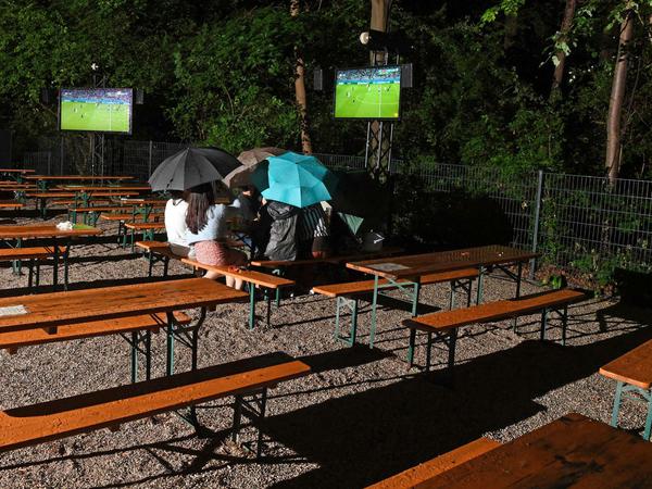 Fans verfolgen das Spiel in einem Biergarten in München bei Regen unter Regenschirmen. 