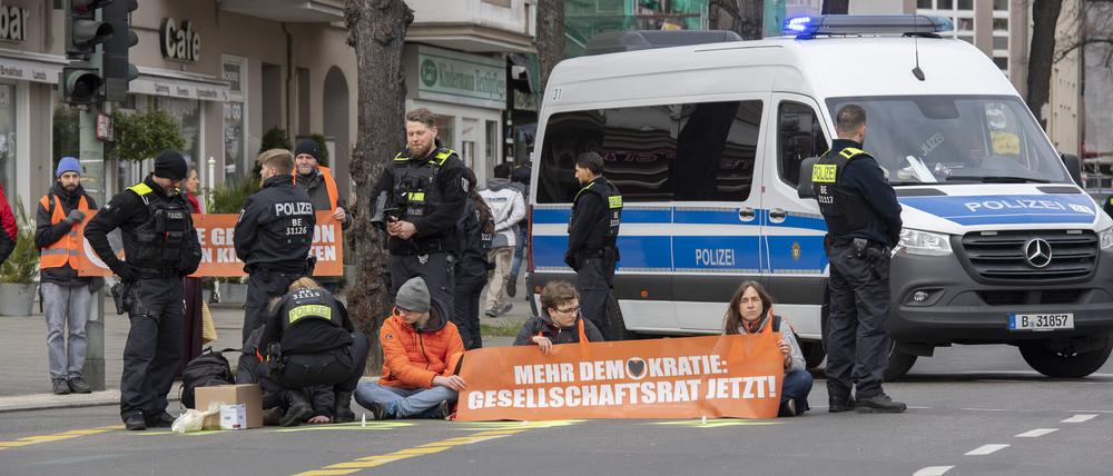 Aktivisten der Gruppe „Letzte Generation“ haben sich auf der Neuen Kantstraße auf einer Kreuzung festgeklebt. Die Polizei sichert den Bereich, während eine Polizistin begonnen hat, die Aktivisten von der Straße zu lösen.