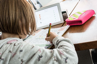 Eine Schülerin sitzt an ihrem Schreibtisch und schreibt in ein Schulbuch, vor ihr steht ein Tablet-Computer. Foto: imago images/Fotostand