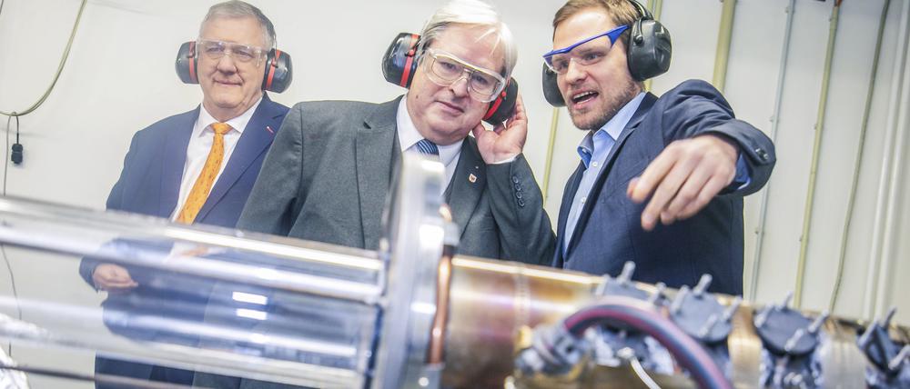 Brandenburgs Wirtschaftsminister Jörg Steinbach (Mitte) lässt sich an der TU Berlin von Tom Tanneberger (r.) zeigen, wie sich Wasserstoff am saubersten und effizientesten verbrennen lässt.