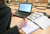 Ein Lehrer sitzt mit einem Schulbuch links und einem Tablet rechts an seinem Platz im Lehrerzimmer. Foto: Felix Kästle/dpa