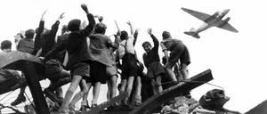 West-Berliner Kinder winken von einem  Trümmerberg  einem US-Transportflugzeug zu, das 1948 während der Blockade Versorgungsgüter bringt.