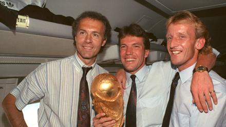 Im Flugzeug beim Rückflug von Rom nach Frankfurt präsentieren DFB-Teamchef Franz Beckenbauer (l), Kapitän und Mittelfeldspieler Lothar Matthäus (M) und Abwehrspieler Andreas Brehme strahlend den zwei Tage zuvor gewonnenen WM-Pokal. 