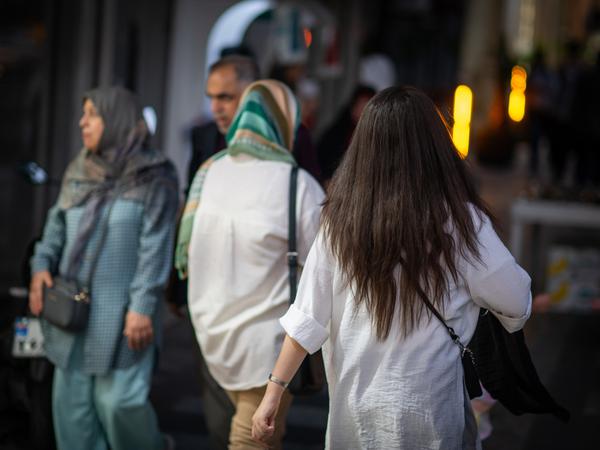 Eine Frau läuft in Teheran mit offenen Haaren am Abend eine Straße entlang, im Hintergrund laufen zwei Frauen mit Kopftuch (Symbolbild).
