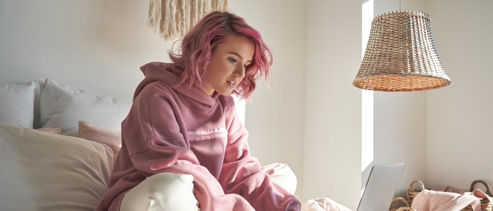 Hipster teen girl school student with pink hair wearing hoodie using laptop computer sitting in bed distance elearning online learning course for exam search remote online classes in bedroom at home.