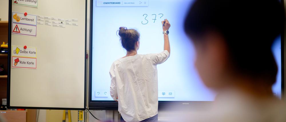 Eine Lehrerin schreibt eine Mathematikaufgabe auf eine digitale Schultafel im Klassenraum einer 4. Grundschulklasse.
