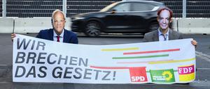 Klimaaktivisten sitzen auf einer Straße und haben sich als Kanzler Olaf Scholz (SPD) und Verkehrsminister Volker Wissing (FDP) verkleidet.