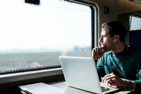 Ein junger Mann sitzt im Zug an einem Laptop und schaut nachdenklich aus dem Fenster. Foto: Getty Images