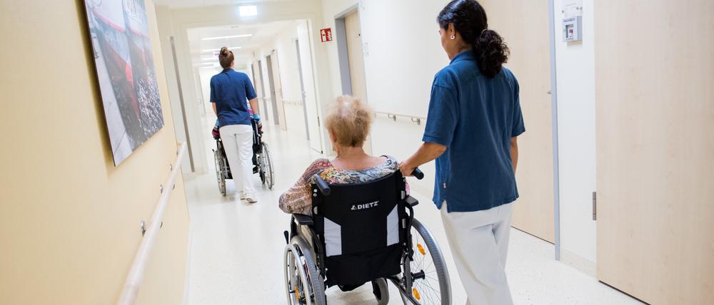 Zwei Pflegerinnen schieben  Patienten mit Rollstühlen durch einen Flur.
