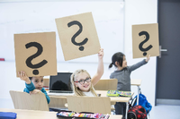 Schüler und Schülerinnen sitzen in einer Klasse und halten Pappschilder mit Fragezeichen hoch. Foto: imago images / Westend61
