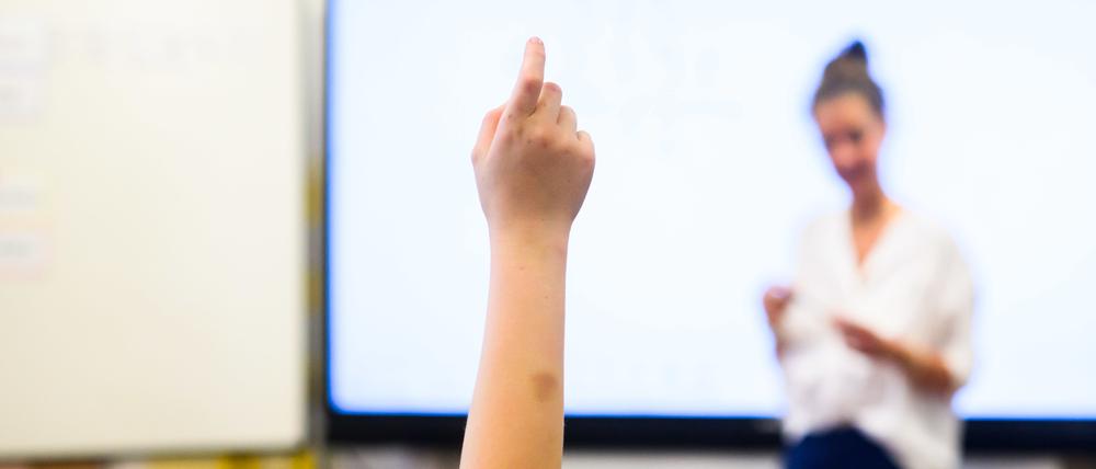 Ein Schüler meldet sich per Handzeichen während eine Lehrerin vor einer digitalen Schultafel steht. 