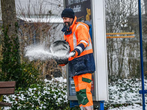 Ein Mitarbeiter des Winterdienstes Heilbronn verteilt Streusalz bei Regen und Schneeglätte auf den Fußweg an einer Bushaltestelle.