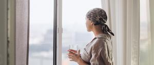 Young unhappy woman with cancer standing near the window and looking at view.