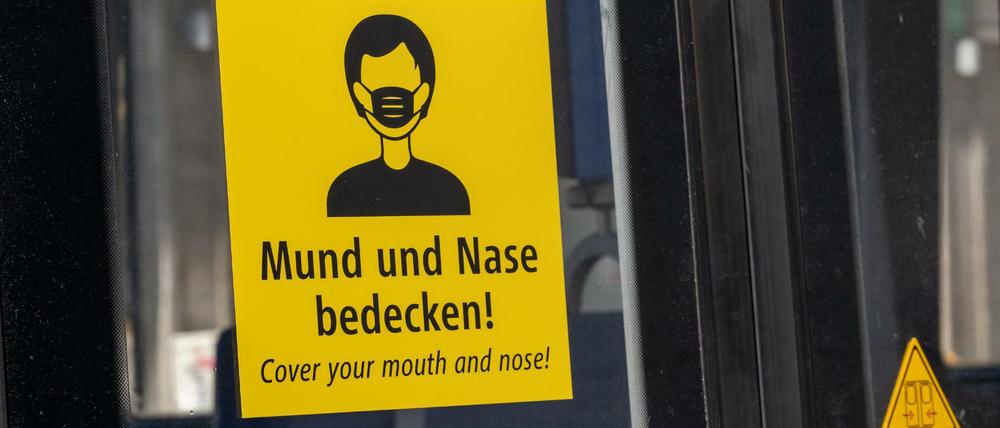 Ein Aufkleber mit der Aufforderung, Mund und Nase zu bedecken, ist in deutscher und englischer Sprache an einer Trambahn in München angebracht. 