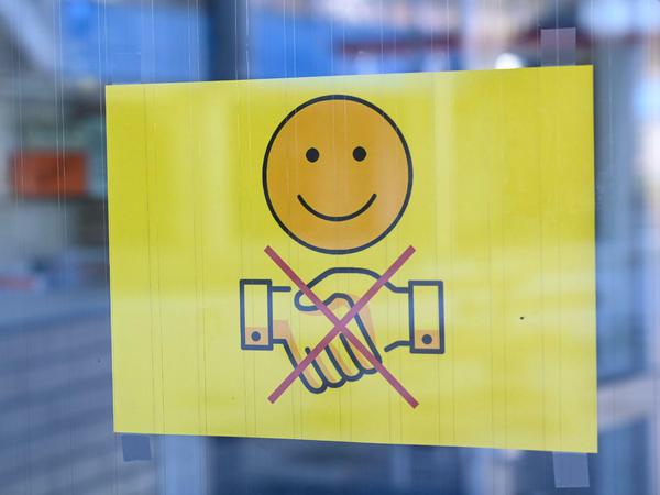 Der Handschlag könnte dauerhaft verschwinden - ein Schild, das ein freundlich lächelndes Gesicht und zwei durchgestrichen Hände, die zum Gruß gereicht werden, zeigt, hängt an der Eingangstür eines Gymnasiums in Baden-Württemberg