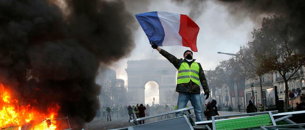 Ein Demonstrant mit einer gelben Weste schwenkt die französische Flagge. Im Hintergrund der Arc de Triomphe.