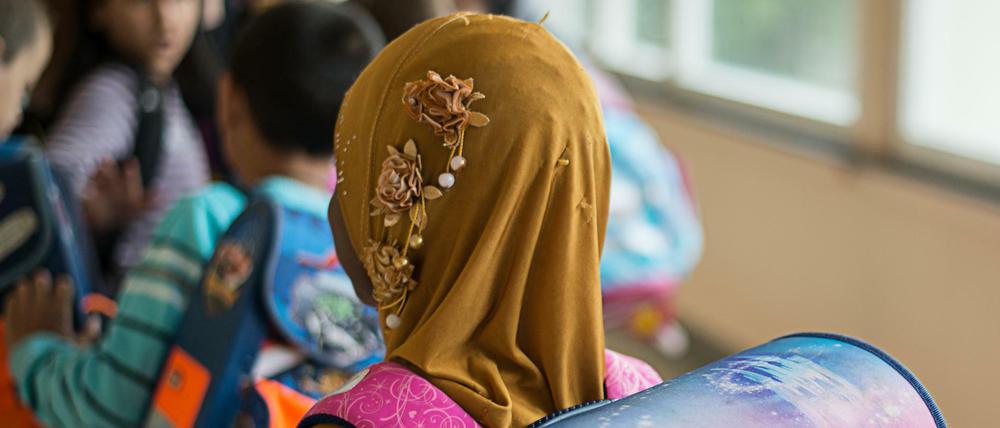Ein Mädchen mit Kopftuch steht in einer Schule vor einem Klassenzimmer. 