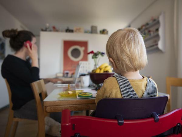 Eine Frau sitzt im Homeoffice an ihrem Laptop und telefoniert, während ihr Kind neben ihr sitzt.