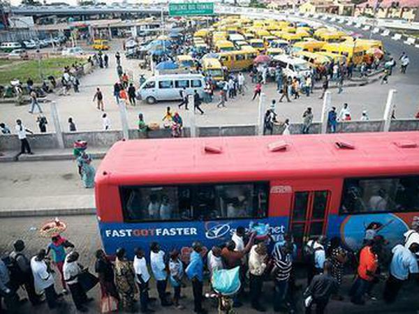 In Lagos stehen Menschen Schlange, um in einen Bus einzusteigen. Im Hintergrund sind Sammeltaxis zu sehen.