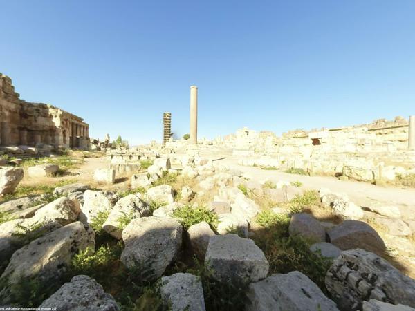 Ein antikes Ruinenfeld mit einer stehenden römischen Säule und einem eingerüsteten Komplex im Hntergrund.