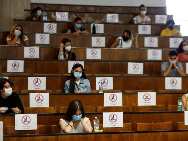 Studierende sitzen mit Mundschutz in einem Hörsaal. Zwischen ihnen sind jeweils mehrere Plätze mit einem Verbotshinweis gesperrt.
